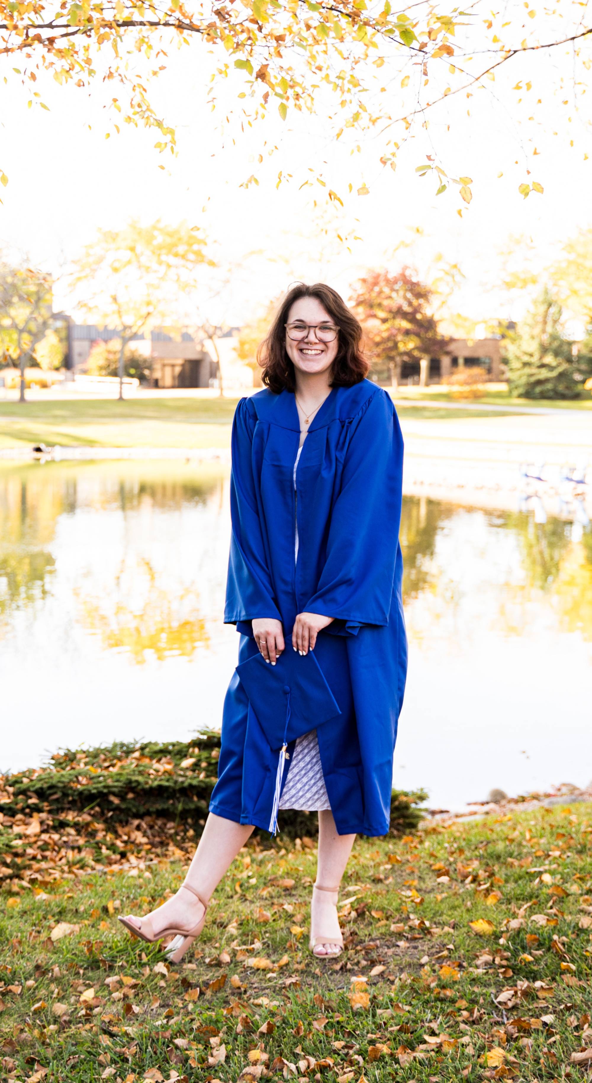 Nichole stands in front of a pond on GVSU's campus in the Fall. She's in her blue graduation gowns, smiling at the camera.